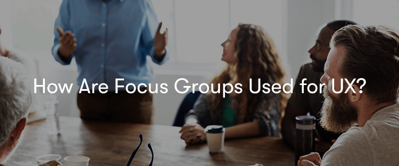 A group of people sat around a table having a discussion with one man standing with the words 'How Are Focus Groups Used for UX?' in front.