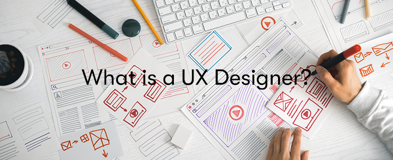 Pieces of paper on a desk with someone drawing UX layouts on them with the text What is a UX Designer? in front